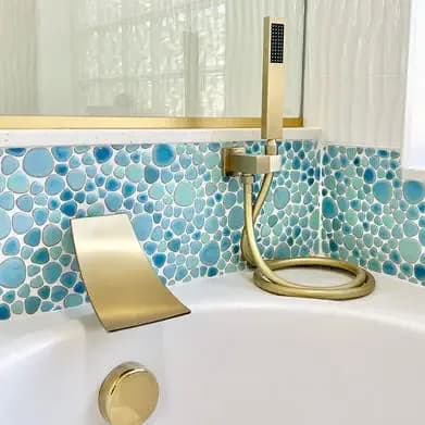 A close-up of a brushed gold tub filler against a blue pebble tile surround