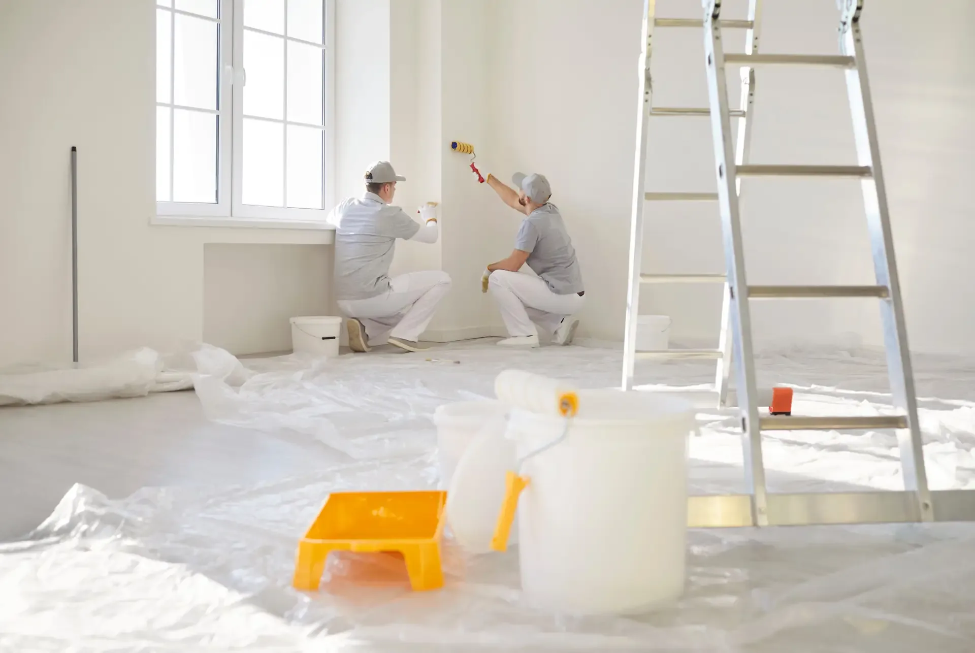 Two painters working on the walls of an empty interior room with a ladder and drop cloths